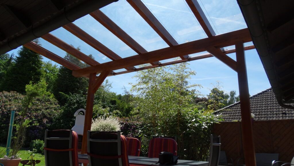 Terrasse mit Glasdach, Gartenmöbeln und üppigem Garten im Hintergrund bei sonnigem Wetter.