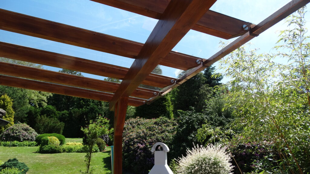 Idyllischer Garten mit Holzpergola, weißem Kamin und üppigem Grün unter klarem, blauem Himmel.