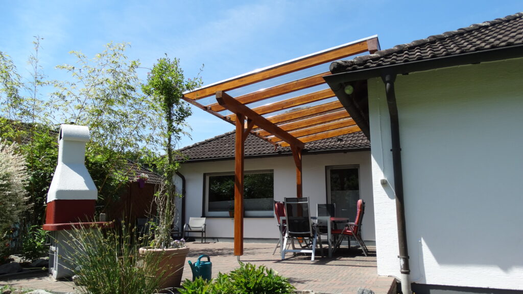 Terrasse mit Tisch, Stühlen, Pergola und Grill, umgeben von Pflanzen, unter klarem, blauem Himmel.