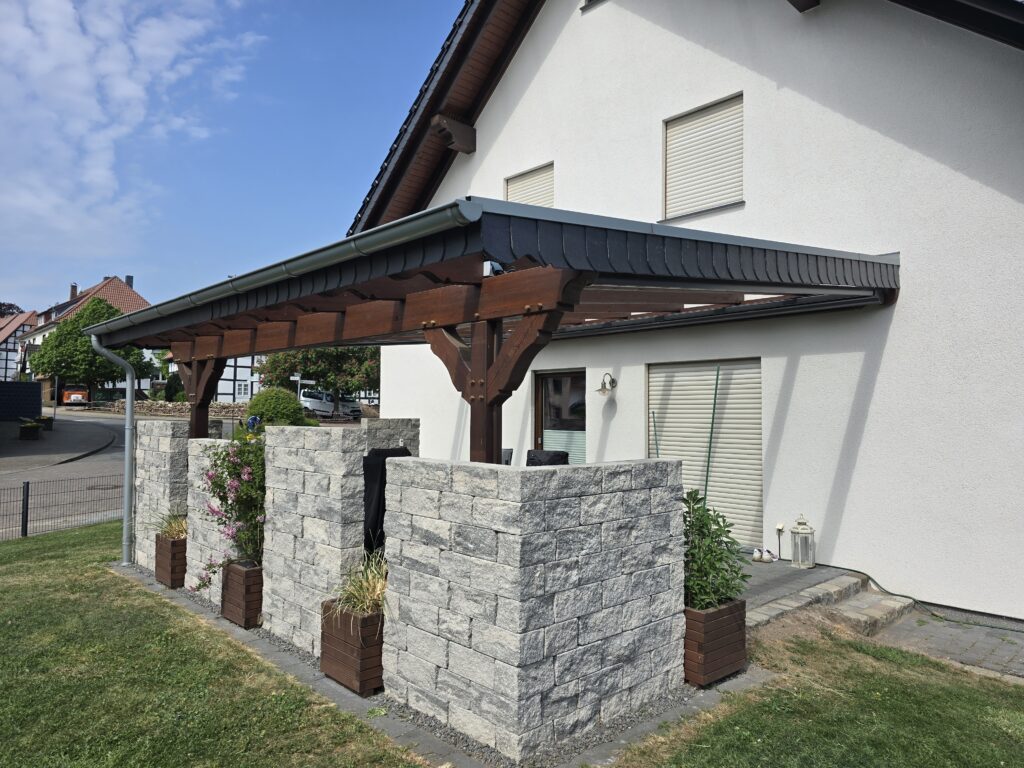 Modernes Haus mit weißer Fassade, überdachter Terrasse und Pflanzen in Holzkübeln, bei sonnigem Wetter.