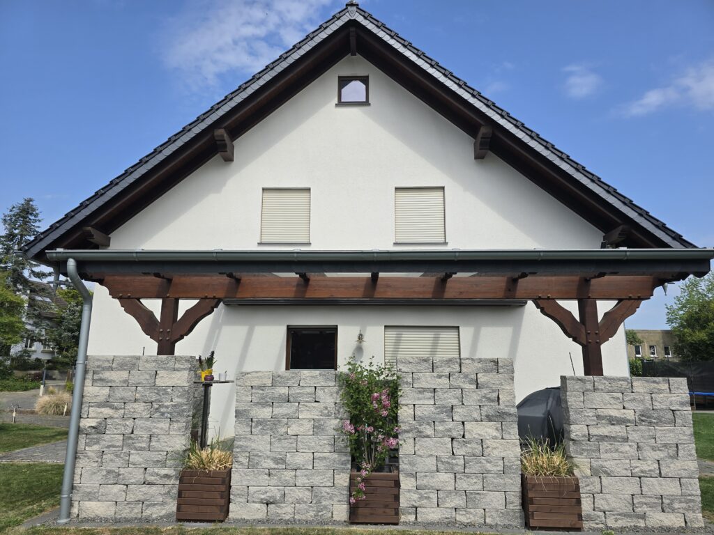 Ein gepflegter Garten mit weißen Hauswänden, grauen Steinmauern, Pflanzenkästen und einer Pergola.