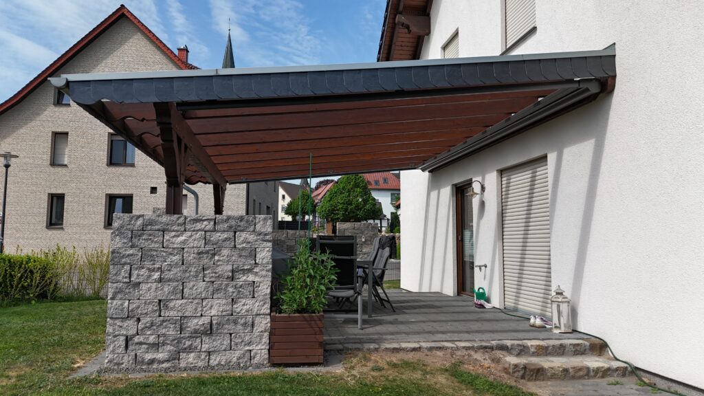 Moderne Terrasse mit Holzbalken, Gartenmöbeln und Steinblock, umgeben von gepflegtem Rasen.