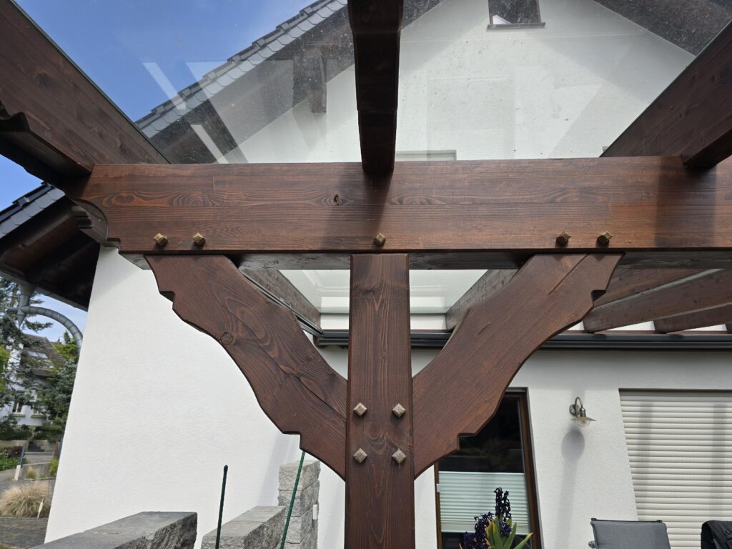 Moderne Holzpergola vor weißem Haus, umgeben von Gartenmöbeln und Pflanzen unter blauem Himmel.