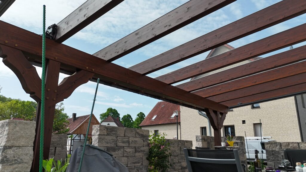 Terrasse mit dunkler Holzpergola, Gartenmöbeln und Pflanzen, umgeben von grauen Steinmauern.