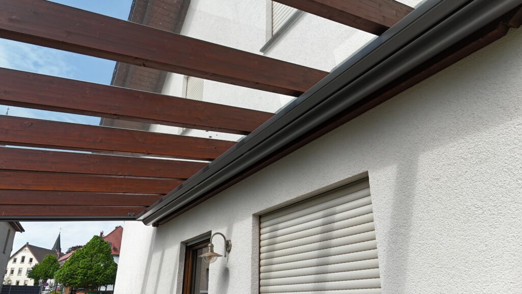 Außenansicht eines Hauses mit Holzpergola, weißer Wand, geschlossenem Rollladen und klarer Himmel.