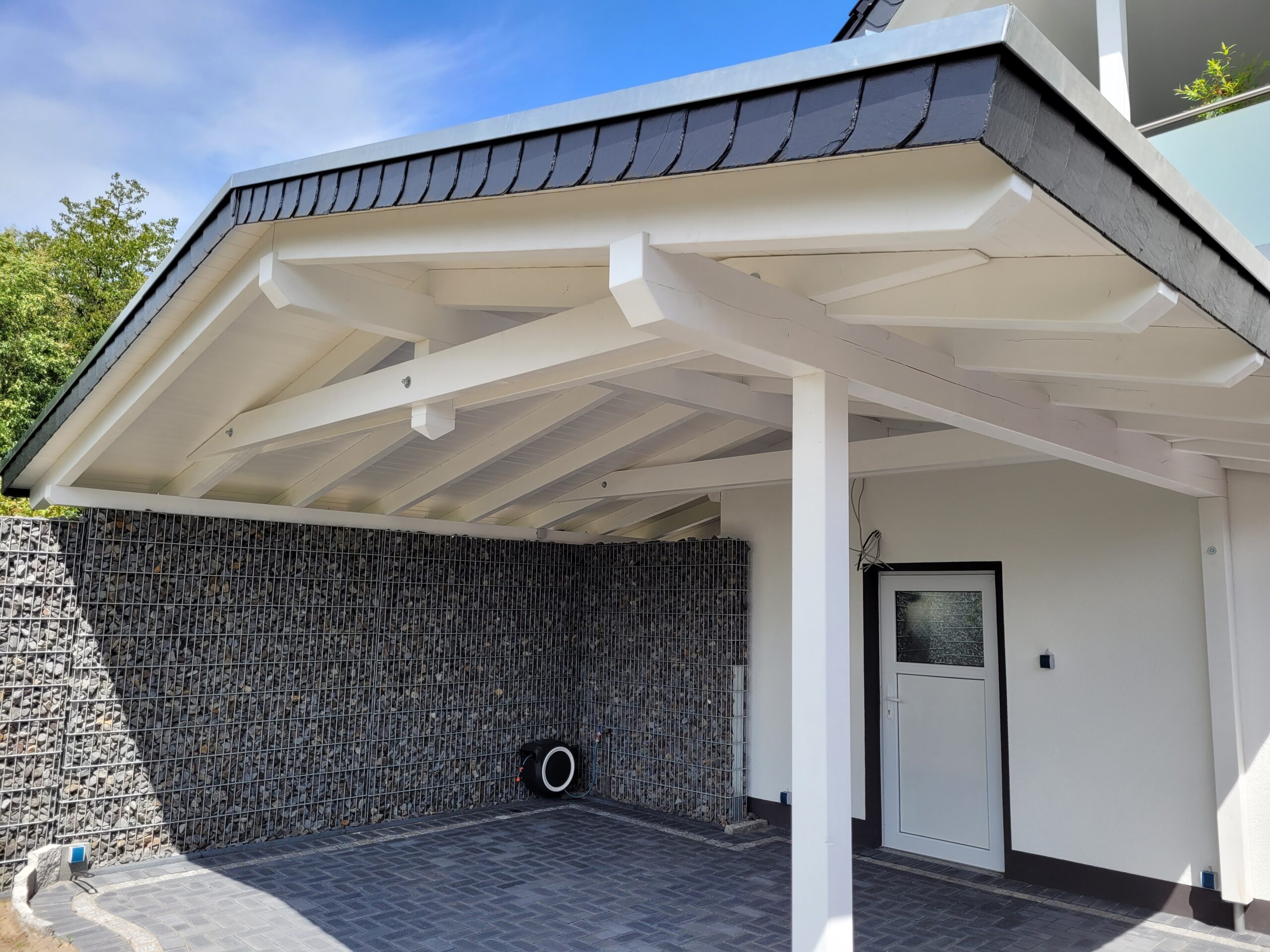 Moderner Carport mit weißem Dach, Holzbalken, Gabionenwand und weißer Tür, sonniger Tag.