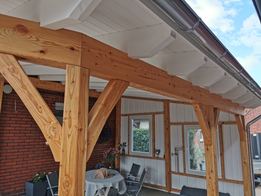 Überdachte Terrasse mit Holzbalken, Tisch und Pflanzen; einladend und gemütlich, ideal zum Entspannen.