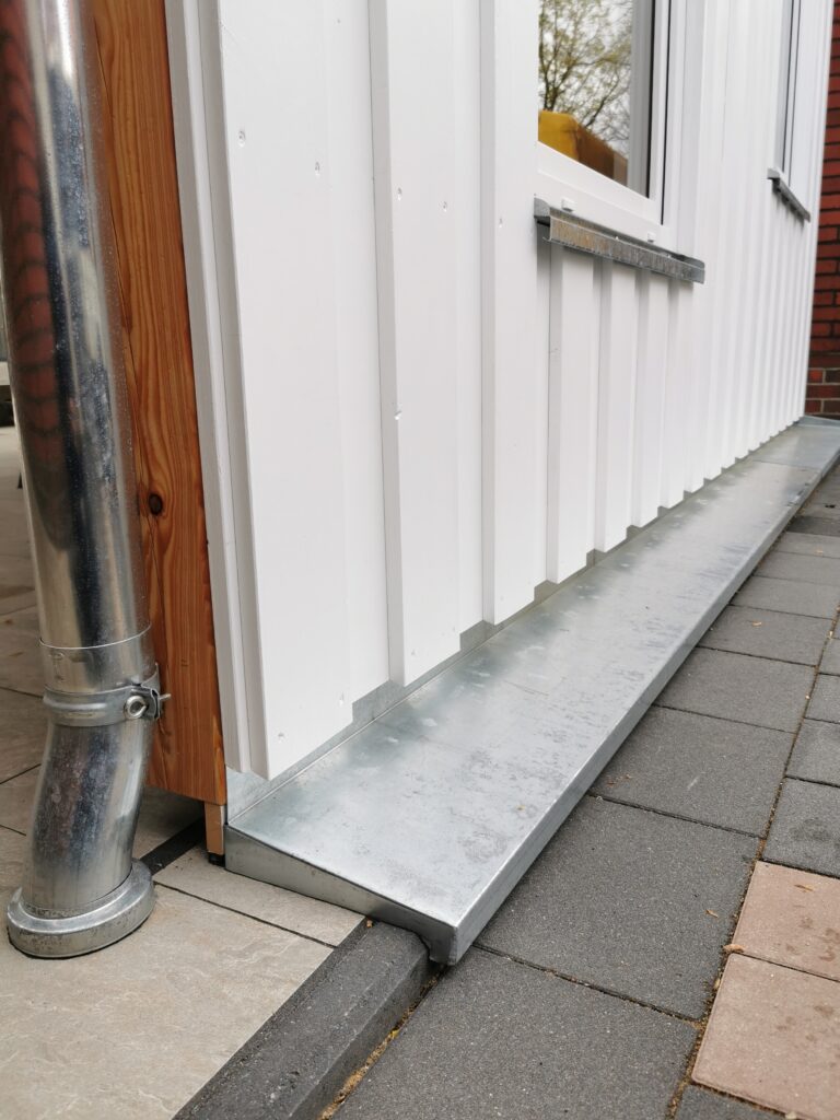 Moderne Fassade mit weißer, geriffelter Wand, silbernem Fallrohr und grauen Pflastersteinen.