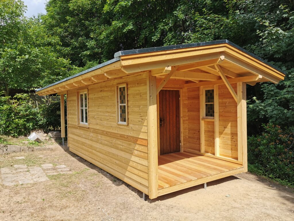 Ein kleines, neu gebautes Holzhaus steht in einem grünen Garten, umgeben von Bäumen und Sträuchern.