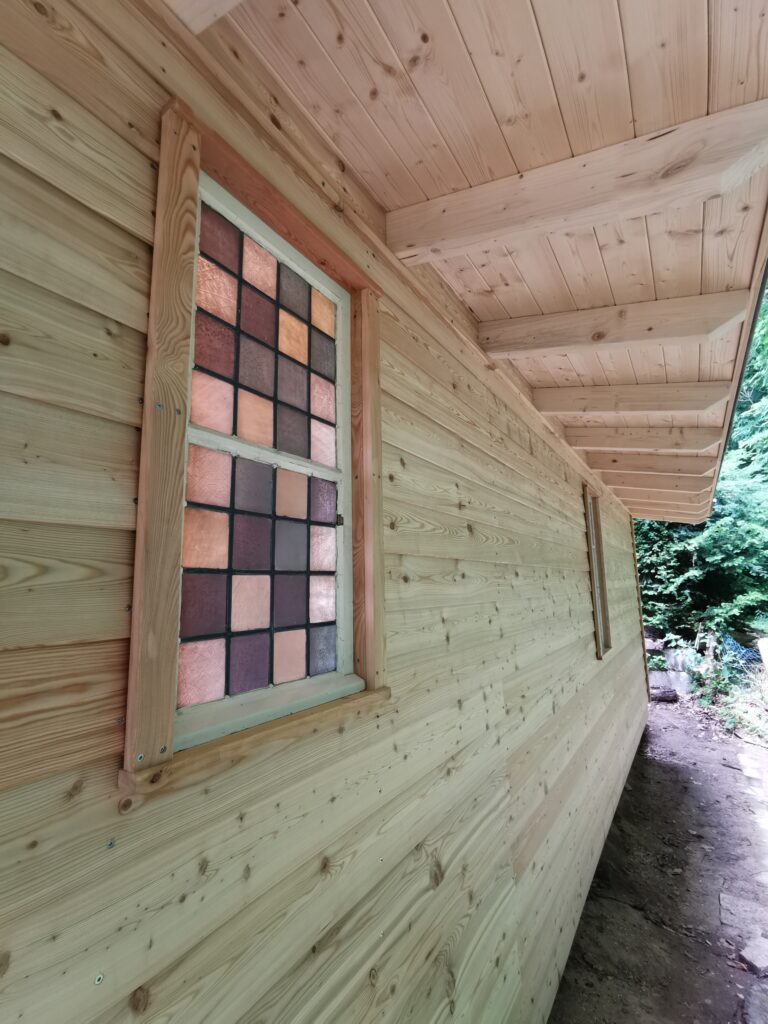 Holzfassade mit Buntglasfenster in Rosa, Lila und Orange, natürliche Maserungen und ruhige Atmosphäre.