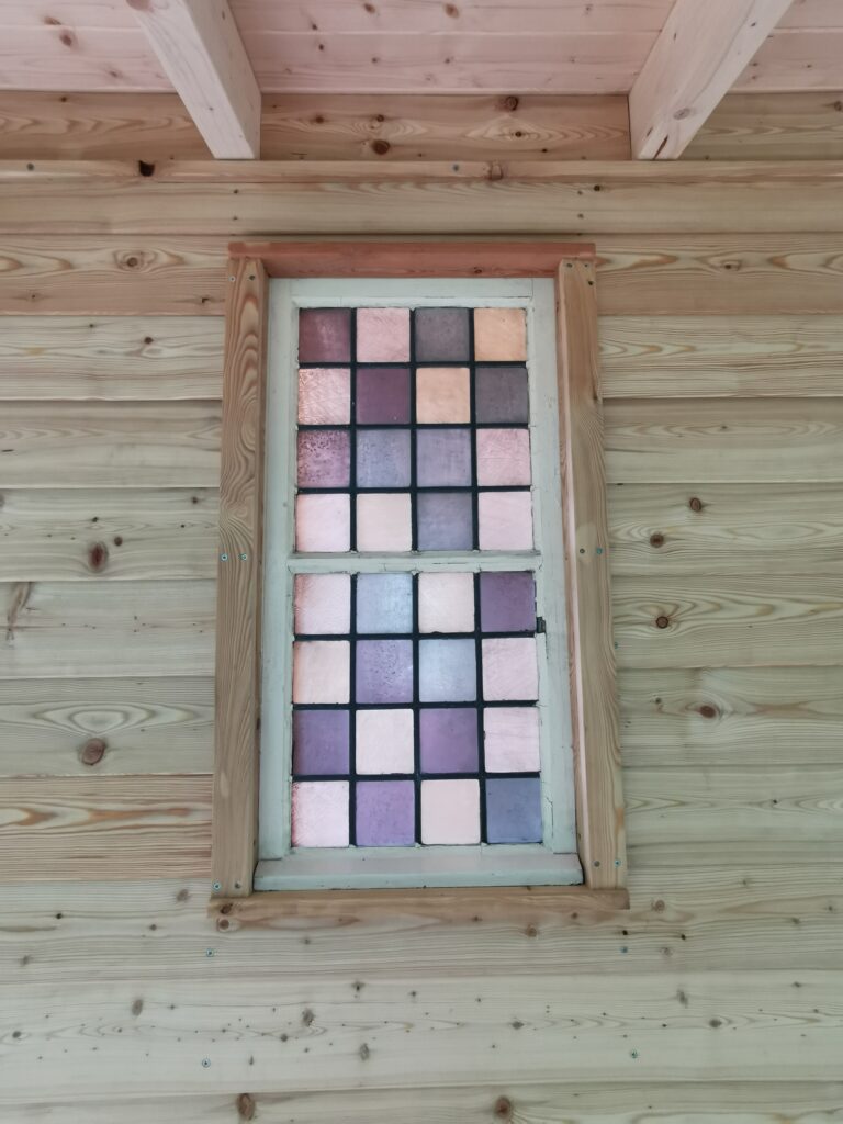 Ein Fenster mit bunten Glasquadraten in Pastelltönen in einem rustikalen Holzrahmen.
