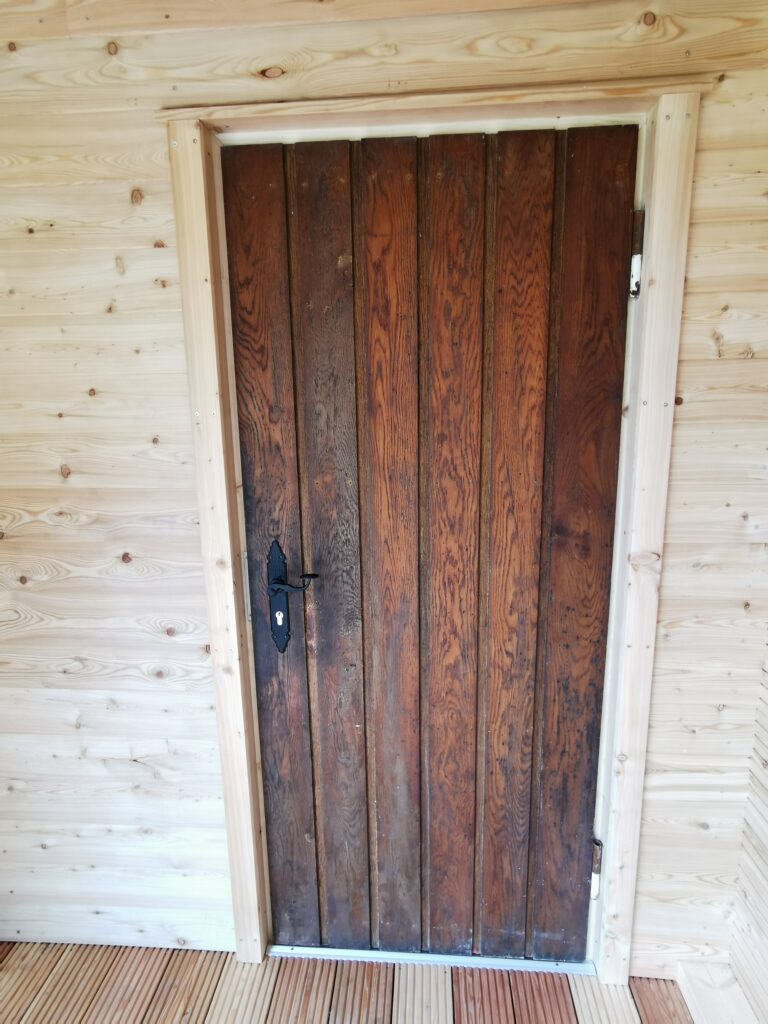 Rustikale Holztür in heller Holzrahmenwand, mit dunklem Türgriff und Schlüsselloch links.