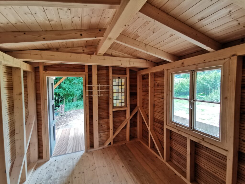 Innenraum einer Holzhütte mit Holzwänden, Boden und Decke, Fenster und Tür mit Blick auf grüne Natur.