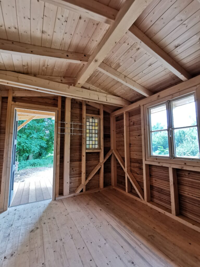 Innenansicht eines Holzhauses mit Holzwänden, Fenster, offener Tür und Blick auf grüne Vegetation.