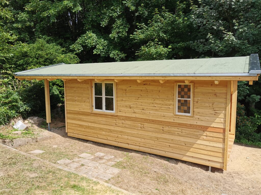 Ein kleines, hölzernes Gartenhaus mit grünem Dach steht in einer grünen, bewaldeten Umgebung.