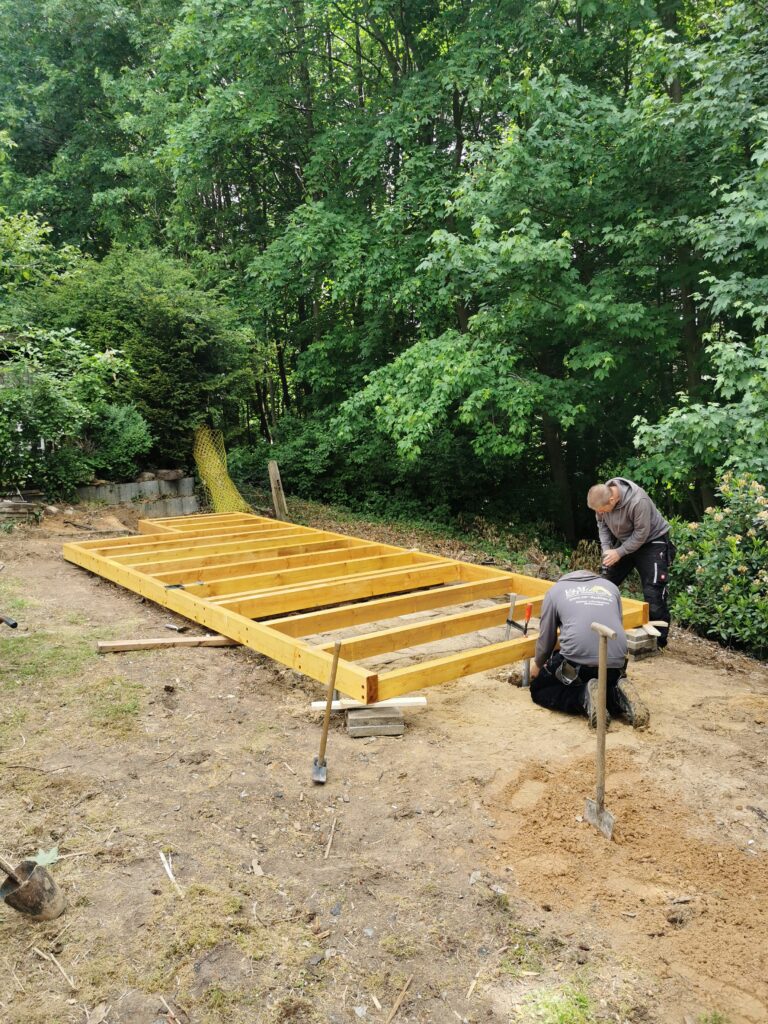 Zwei Personen arbeiten an einem Holzrahmen auf einer Baustelle im Freien, umgeben von grüner Vegetation.