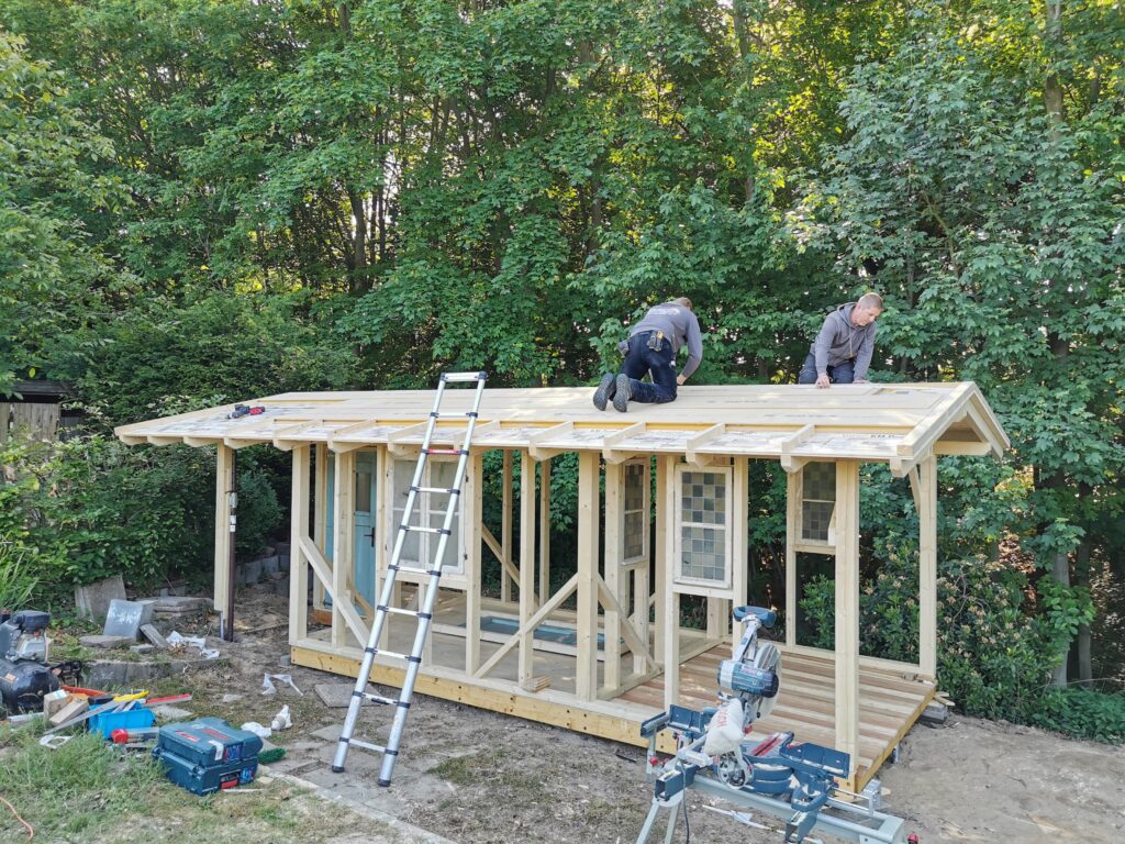 Zwei Personen bauen konzentriert das Dach eines Holzschuppens in einem grünen Garten.