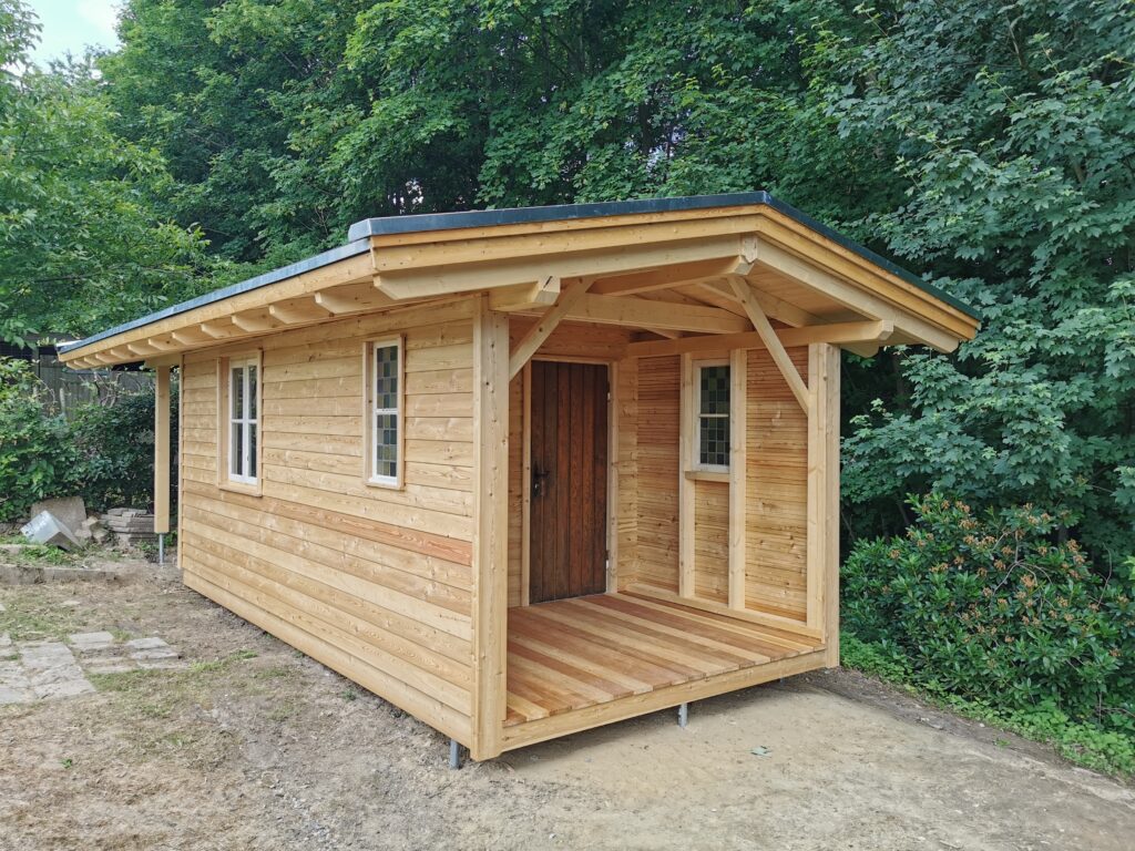 Ein kleines, hölzernes Gartenhaus mit Flachdach, umgeben von grünen Bäumen und Büschen.