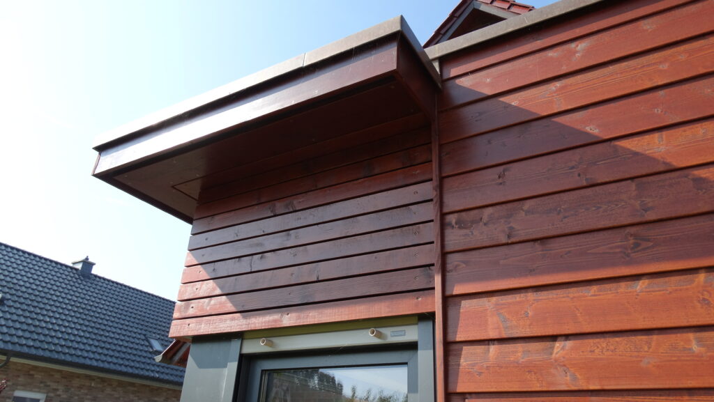 Nahaufnahme einer Holzfassade mit rötlich-braunen Latten, teils sichtbares Fenster und blaues Dach im Hintergrund.