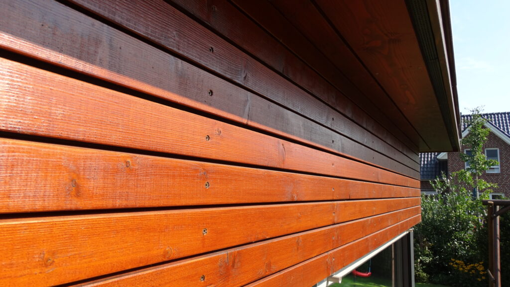Seitenansicht einer Holzfassade mit Garten im Hintergrund, sonniges Wetter und einladende Atmosphäre.