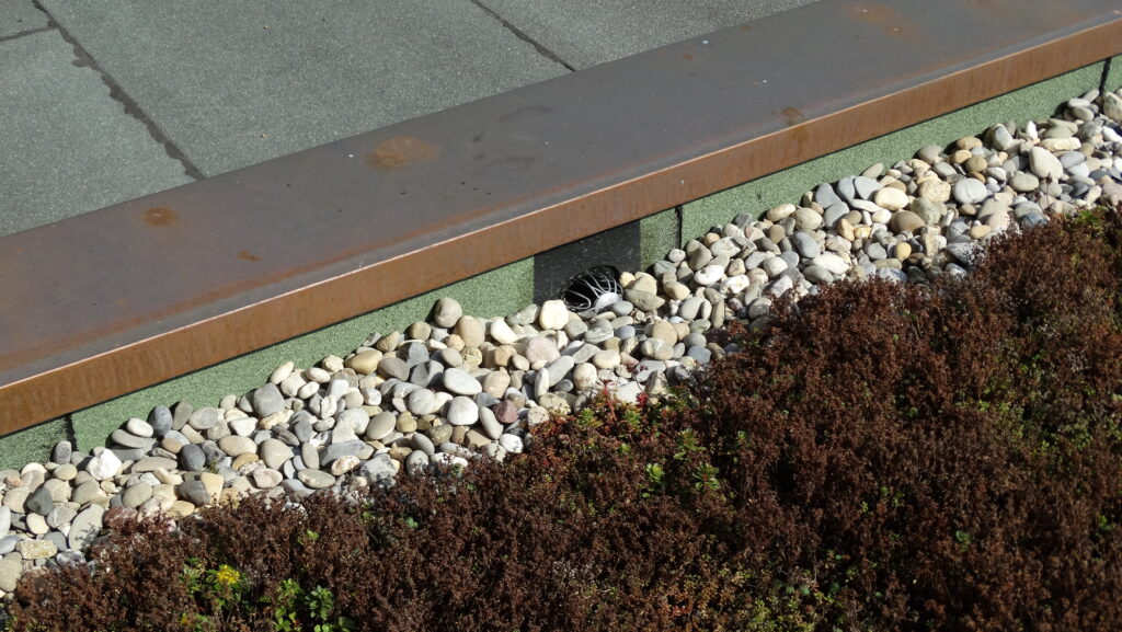 Rostiger Metallrand mit Kieselsteinen und Pflanzen, urbanes Design mit natürlichem Flair.