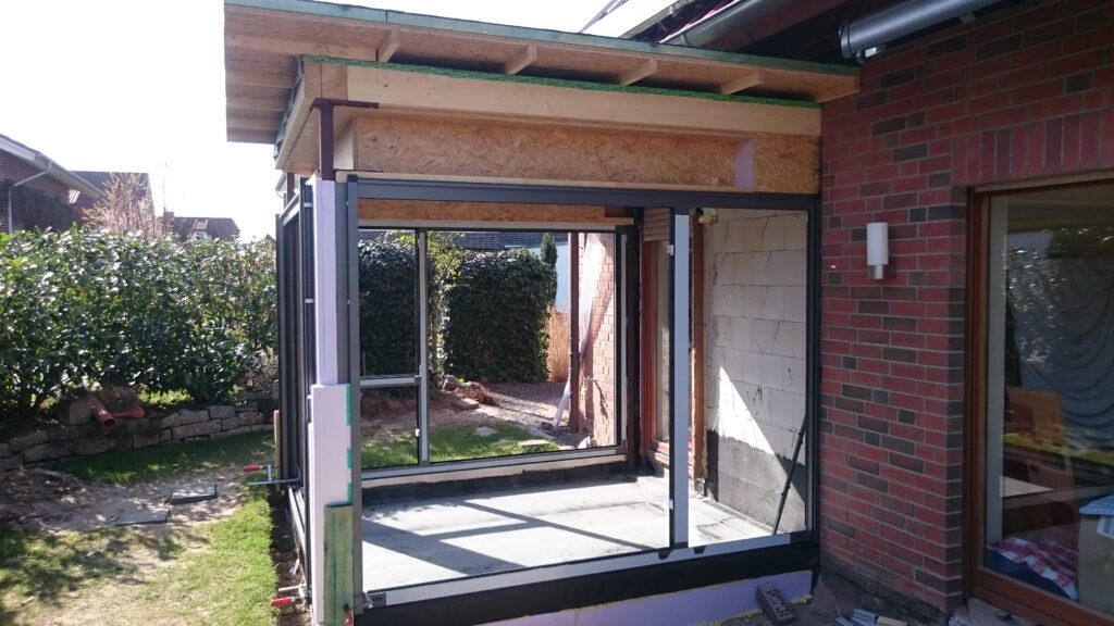 Baustelle: Anbau mit Glaswänden und Holzdach an Ziegelhaus, umgeben von Garten mit Rasen und Hecken.