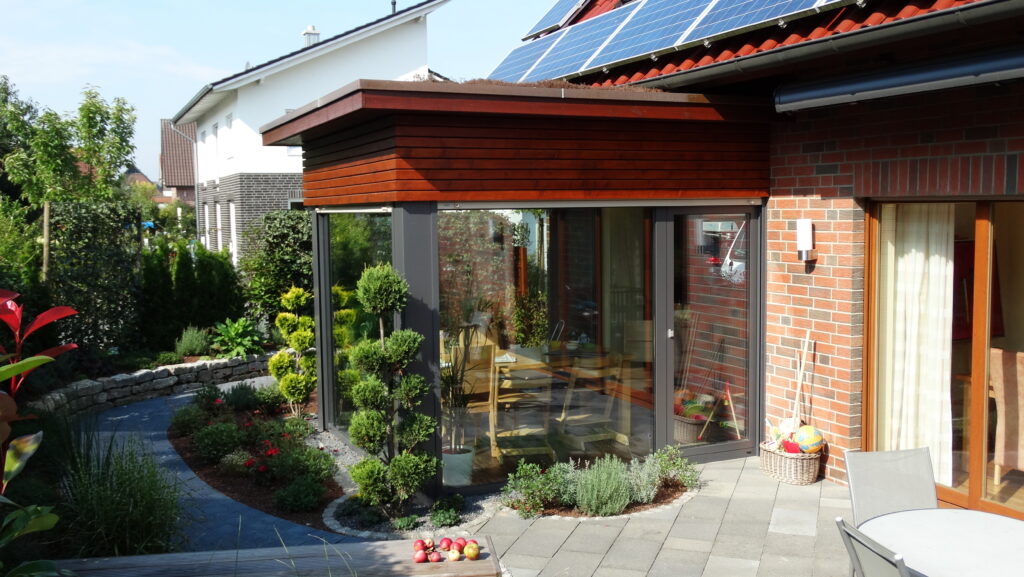 Moderne Hausansicht mit Wintergarten, großen Glasfenstern, Solarpaneelen und gepflegtem Garten.