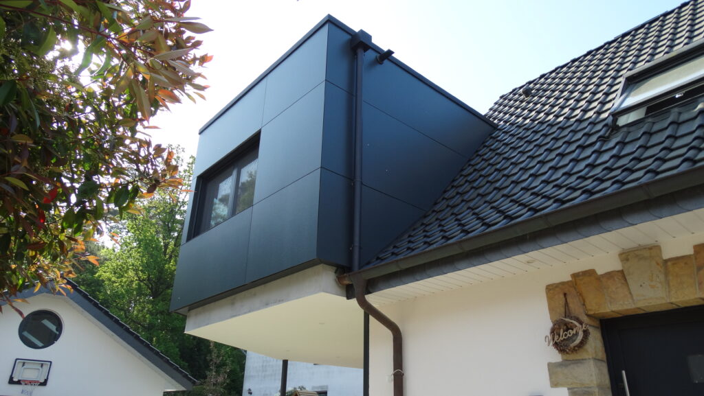 Moderner, kubischer Anbau mit dunkler Fassade neben traditionellem Hausdach und Baum mit bunten Blättern.