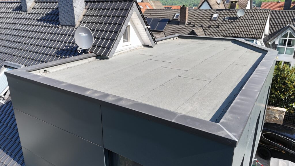 Flachdach eines modernen Gebäudes neben Ziegeldächern mit Satellitenschüsseln an einem sonnigen Tag.