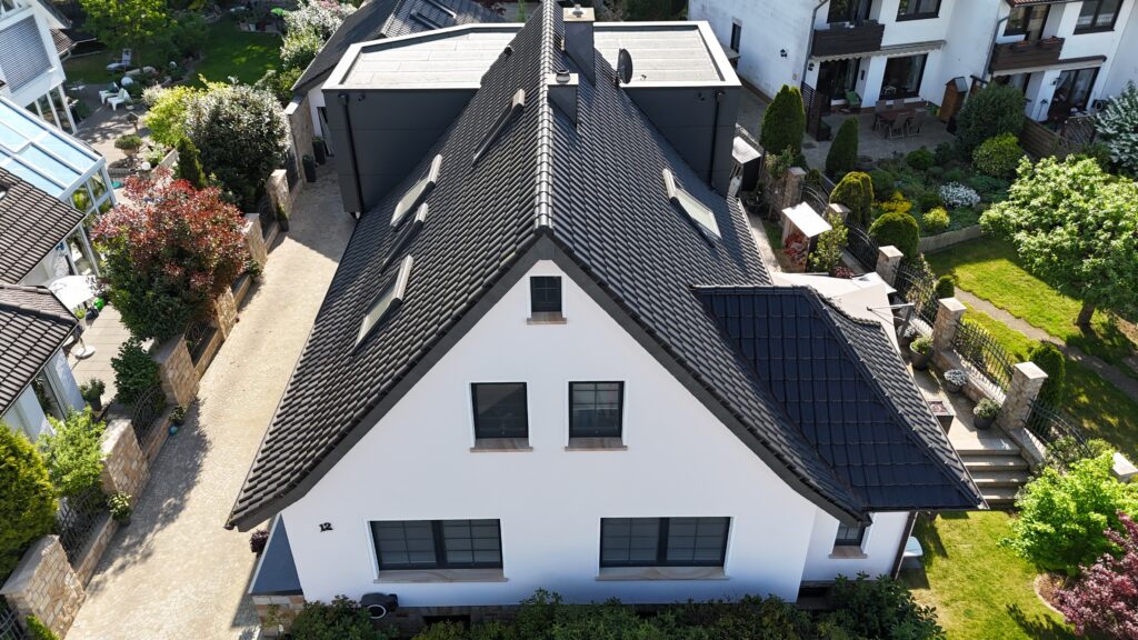 Luftaufnahme eines modernen weißen Hauses mit dunklem Schrägdach, umgeben von gepflegtem Garten.