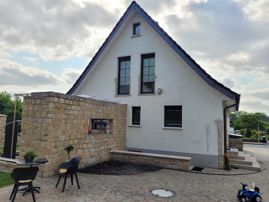 Moderne Hausfassade mit Satteldach, Terrasse mit Stühlen, Tisch und Pflanzen, blaues Spielzeugauto.