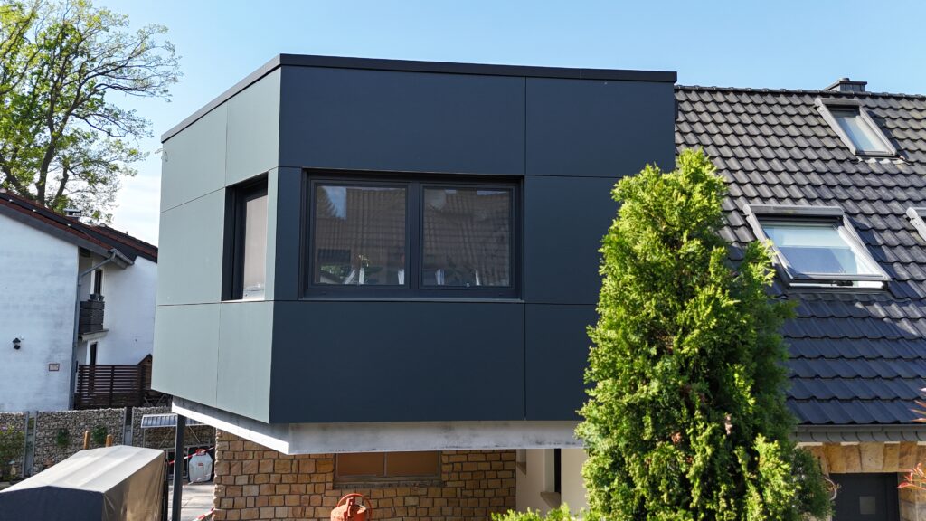 Moderner Anbau mit großen Fenstern an einem Haus, umgeben von Bäumen und blauem Himmel.