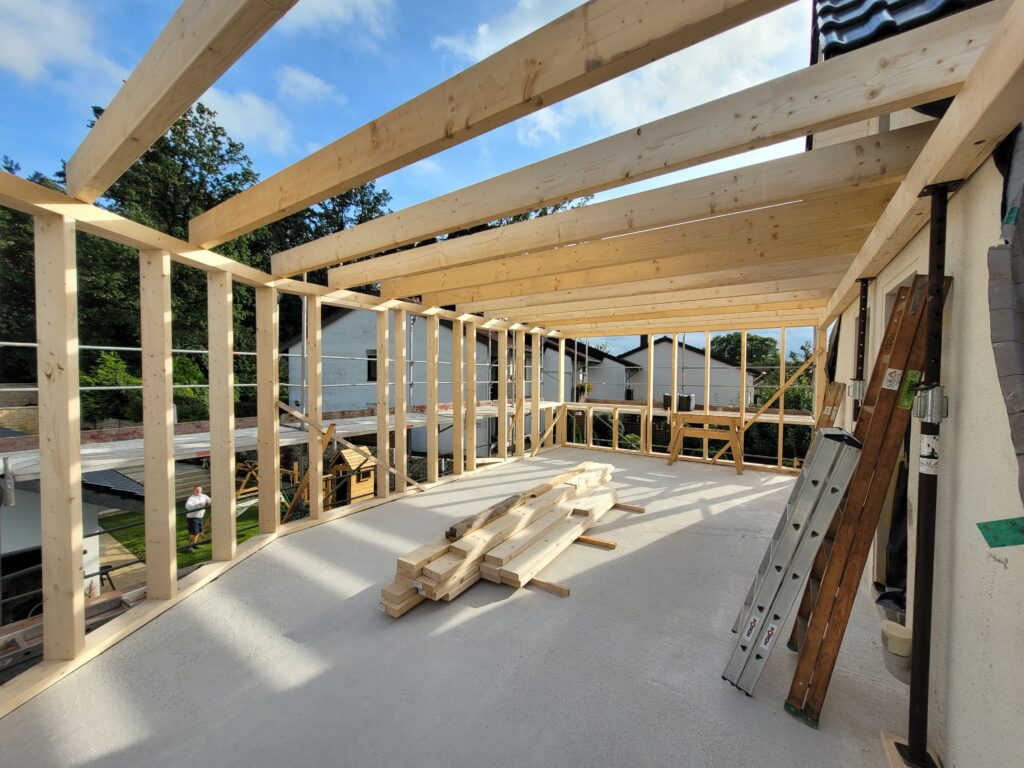 Holzrahmenbau auf Baustelle mit Leiter, Holzlatten und blauem Himmel in ländlicher Umgebung.