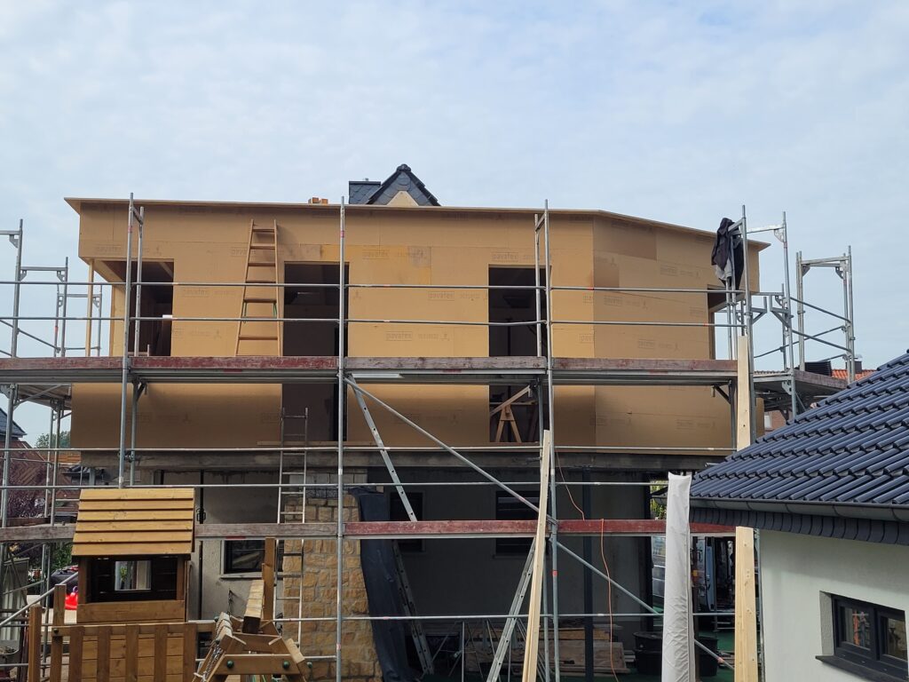 Eine Baustelle mit einem zweistöckigen Rohbau, umgeben von Gerüsten, und einem kleinen Holzspielhaus.