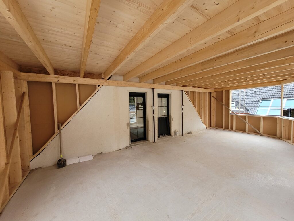 Unfertiger Dachgeschossraum mit Holzkonstruktion, Glastüren, Fenster und Baumaterialien auf Betonboden.