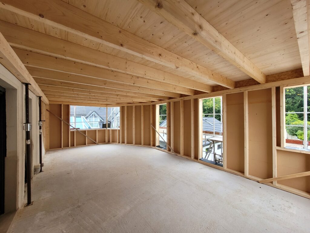 Innenraum eines im Bau befindlichen Hauses mit Holzkonstruktionen und Betonboden, ohne Personen.