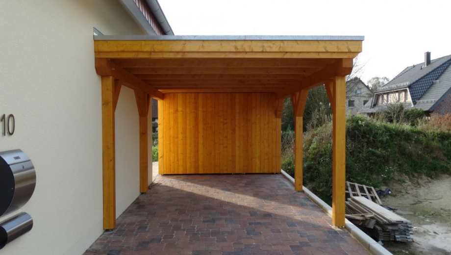 Ein hölzerner Carport an einem cremefarbenen Haus, umgeben von Pflastersteinen und grüner Vegetation.