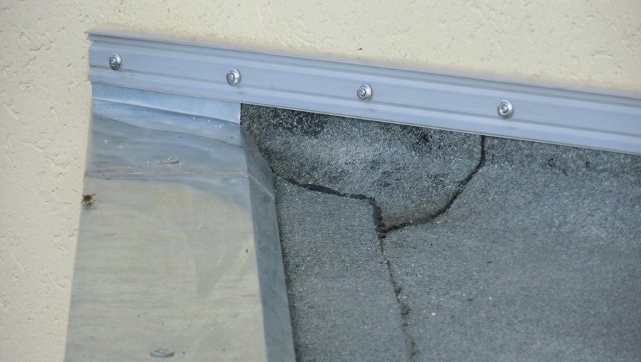 Dachkante mit silberner Metallverkleidung, grauer Dachfläche mit Rissen und cremefarbener Wand.