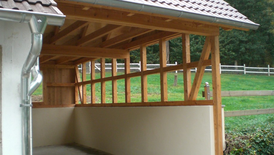 Offene Holzkonstruktion mit Ziegeldach, Regenrinne und betoniertem Boden, umgeben von Rasen und Zaun.
