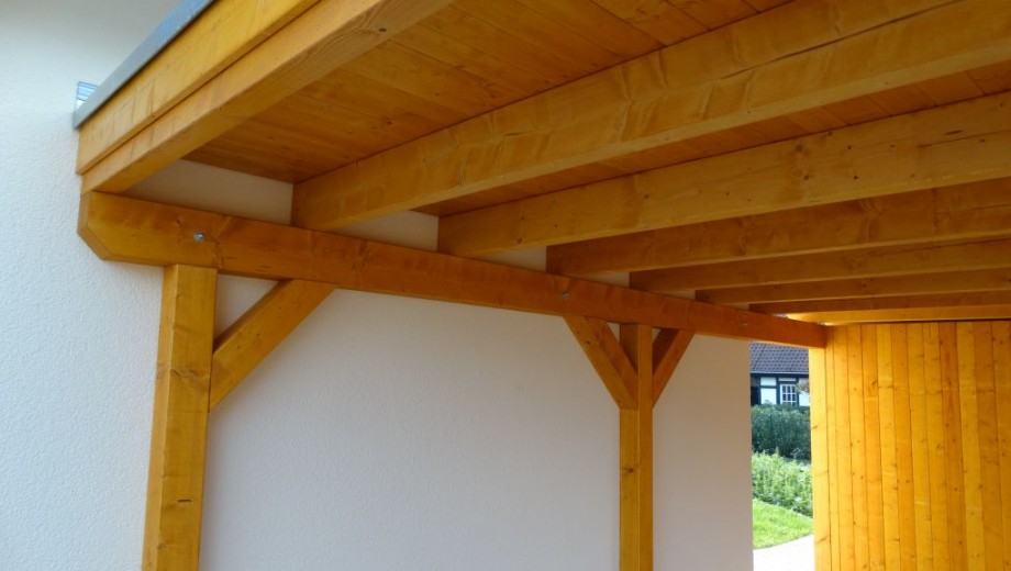 Überdachte Holzstruktur an Hauswand, umgeben von Garten; warme, einladende Atmosphäre.