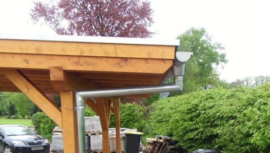 Holz-Carport mit Metallregenrinne, geparktem Auto und gestapelten Materialien im grünen Garten.