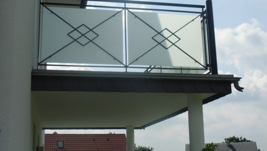 Ein Balkon mit Glasgeländer an einem weißen Gebäude, rotes Ziegeldach und bewölkter Himmel im Hintergrund.