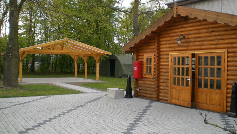 Eine rustikale Holzhütte mit Überdachung und grünem Zelt in einer bewaldeten, gepflasterten Umgebung.