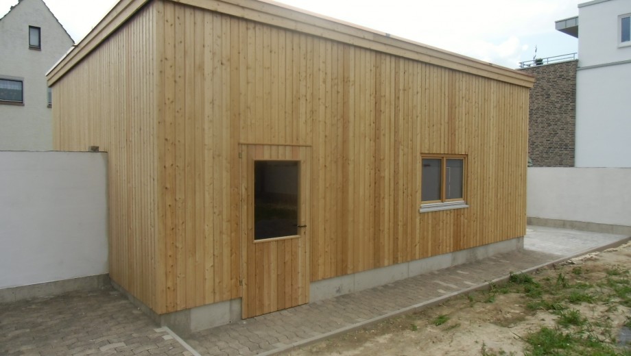 Holzgebäude mit flachem Dach, Tür und Fenster, umgeben von grauen Pflastersteinen und weißem Zaun.