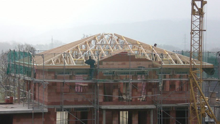 Baustelle mit Rohbau eines Hauses, zwei Arbeiter am hölzernen Dachstuhl, Kran im Hintergrund.