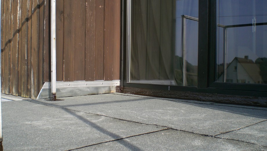 Ecke eines modernen Gebäudes mit Holzwand, großer Glasfläche und grauem Steinboden, bei Tageslicht.