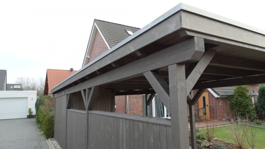 Ein grauer Holz-Carport steht vor einem roten Ziegelhaus, umgeben von Pflanzen und einem gepflasterten Weg.