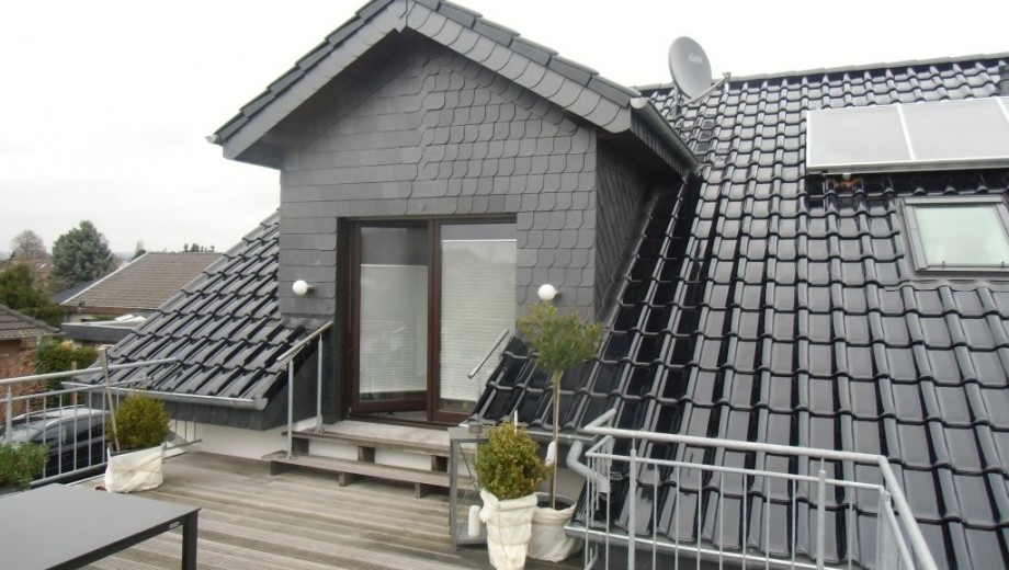 Moderne Dachterrasse mit Holzdielen, dunklem Dach, Glastür, Metallgeländer und grünen Pflanzgefäßen.