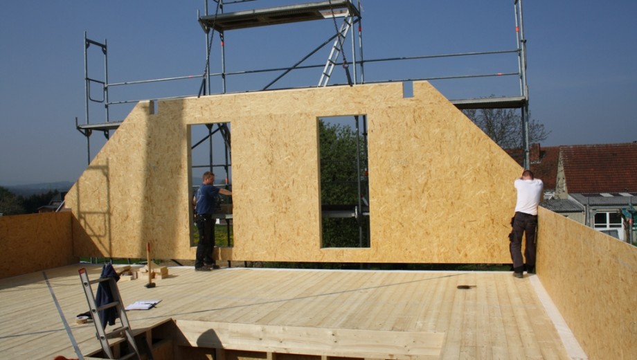 Zwei Personen arbeiten an einer Holzwand auf einer sonnigen Baustelle, umgeben von einem Gerüst.