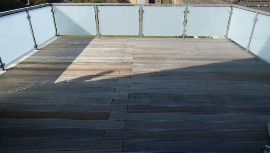 Eine Terrasse mit Holzboden und einem Geländer aus Metall und milchigem Glas, beleuchtet von Sonnenlicht.